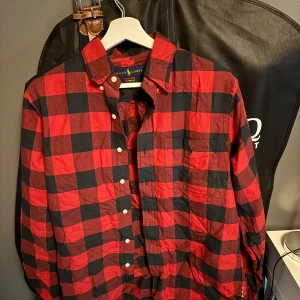 Röd och svart rutig skjorta Ralph Lauren - Snygg röd och svart rutig skjorta från Ralph Lauren i klassisk passform. Skjortan har knappar framtill, bröstficka och klassisk krage. Perfekt för dig som gillar en tidlös och avslappnad stil.