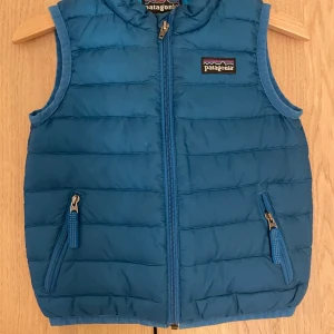 Blå dunväst från Patagonia 18M - Snygg blå dunväst från Patagonia med quiltad design och dragkedja framtill. Västen har två fickor med dragkedja och klassisk Patagonia-logga på bröstet. Perfekt för lager-på-lager under kyliga dagar.