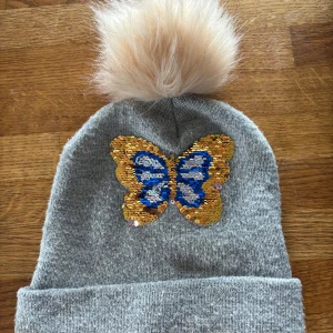 Grå mössa med paljettfjäril och pälsboll - Grå stickad mössa med stor fluffig beige pälsboll på toppen. Framtill finns en paljettbroderad fjäril i guld, rosa och blått som skiftar färg beroende på hur du vänder paljetterna. Perfekt accessoar för att sticka ut under kalla dagar.