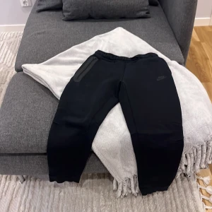 Svarta Nike mjukisbyxor med dragkedja - Svarta mjukisbyxor från Nike med diskret logga på låret och elastisk midja med snörning. Ena fickan har en snygg dragkedja med gummidetalj. Byxorna är i mjukt material och har ribbade muddar vid bensluten för en skön passform. Köpt från Nike butik