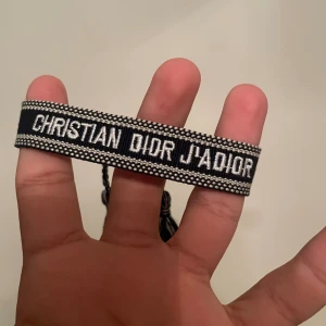 Christian Dior J'Adior armband - Snyggt armband från Christian Dior med texten 'J'Adior' broderad i vitt på svart bakgrund. Armbandet har justerbar knytning och dekorativa tofsar i svart och vitt. Perfekt accessoar för att lyfta din outfit med en lyxig touch.