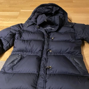 Mörkblå dunjacka från Boomerang XL - Säljer en mörkblå dunjacka från Boomerang i storlek XL. Jackan har huva, stora fickor och stängs med både knappar och dragkedja. Klassisk pufferstil med broderad logga på ärmen. Perfekt för kalla dagar när du vill hålla värmen och ändå se snygg ut.