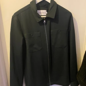Svart overshirt från Calvin Klein - Stilren svart overshirt från Calvin Klein med klassisk krage, två bröstfickor med knapp och dragkedja framtill. Jackan har lång ärm och diskret logga på ärmen. Perfekt för lager-på-lager och enkel att matcha. Perfekt för hösten