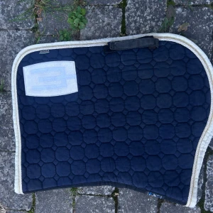 equline - Bilden visar en sadelpad för häst, inte en elektronikprodukt. Produkten är mörkblå med vit kant och verkar vara i använt men gott skick. Inga synliga defekter eller större slitage. Passar bra för ryttare som söker en funktionell sadelpad.