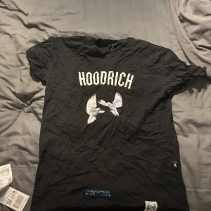 Svart HOODRICH t-shirt med duvor - Svart t-shirt från HOODRICH med tryck av vita duvor och text både fram och bak. Klassisk passform med korta ärmar och rund hals. Snygg streetwear-stil med coola detaljer och tydlig logga framtill. Perfekt för dig som gillar statement-plagg.