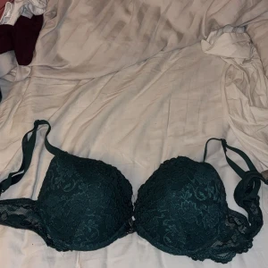 Mörkgrön spets-bh med rosett - Snygg mörkgrön bh med vadderade kupor och vacker spets över hela framsidan. Dekorerad med en liten svart rosett och ett litet smycke mellan kuporna. Axelbanden är justerbara och sidorna har genomskinlig spets. Perfekt för dig som gillar detaljer och en elegant look.