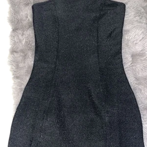 Glittrig svart tubklänning med slits - Snygg svart tubklänning med glittrigt tyg och en liten slits framtill. Klänningen är tight och kort, perfekt för dig som vill sticka ut. Enkel och stilren design utan axelband som är perfekt nu till nyår eller till en utekväll💓