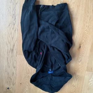 Svart hoodie från Polo Ralph Lauren - Svart hoodie från Polo Ralph Lauren med dragkedja och huva. Klassisk design med liten röd logga broderad på bröstet. Tillverkad i mjuk bomull och har långa ärmar. Perfekt för en chill och stilren look.