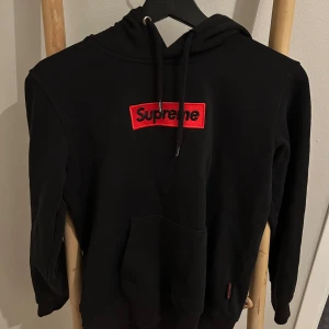 Svart Supreme hoodie - Svart hoodie från Supreme med klassisk röd broderad logga på bröstet. Tröjan har huva med snörning, känguruficka och långa ärmar. Perfekt för dig som gillar streetwear och vill ha en stilren look.