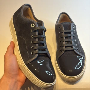 Lanvin sneakers i mocka och lack - Snygga sneakers från Lanvin i mörkgrå mocka med glansig lackad tå och vita gummisulor. Skorna har rund tå, snörning och diskreta silvriga öljetter. Perfekt för dig som gillar en stilren men ändå unik look.