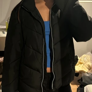 Vinterjacka - Säljer en svart pufferjacka med markerad quiltad design och silverfärgad dragkedja framtill från JDY i stl M. Jackan har en klassisk krage och är långärmad, perfekt för kyligare dagar. Enkel och stilren modell som passar till det mesta.