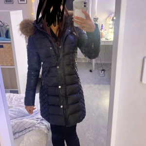 Svart lång Rockandblue dunjacka med päls - Lång svart dunjacka från Rockandblue med snyggt mönster och stor huva med fluffig brun päls. Jackan har Rockandblue-logga på ärmen. Perfekt för kalla vinterdagar. Använd!