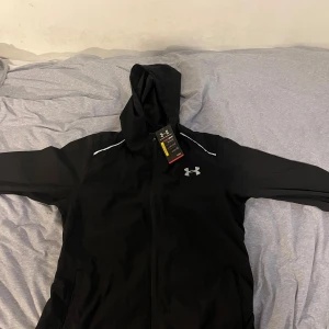 Svart vindjacka från Under Armour L - Svart vindjacka från Under Armour med huva och dragkedja. Jackan har vita detaljer vid axlarna och en stor logga på ryggen. Perfekt för träning eller när du vill ha en sportig look. Tillverkad i lätt polyester som andas. Fick av min vän när han va i Frankrike  men är för stor för mig så därför säljer!