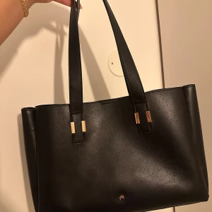 Svart axelväska i skinn från Åhléns - Stilren svart axelväska i skinn från Åhléns med guldiga metalldetaljer på handtagen. Väskan har rymligt huvudfack och stängs med magnetknapp. Perfekt för dig som vill ha en klassisk och snygg väska med plats för allt du behöver.