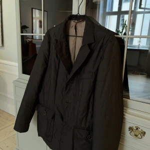 Quiltad kavaj från Morris, brun - Säljer en brun quiltad kavaj från Morris i storlek 48. Jackan har klassisk kavajkrage, två stora fickor med knapp framtill och en bröstficka. Fodrad insida i beige och stängs med knappar. Perfekt för dig som vill ha en stilren och bekväm look.