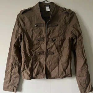 Brun militärinspirerad jacka med knappar - Snygg brun jacka från People Tree med militärvibbar, dekorerad med dubbla rader knappar och axelklaffar. Jackan är figursydd och har långa ärmar samt coola detaljer framtill. Perfekt för dig som vill sticka ut med en unik stil.