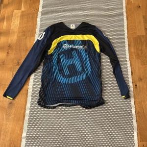 Blå långärmad tröja från Husqvarna - Säljer en blå och svart långärmad tröja från Husqvarna med gula detaljer och coolt randigt mönster. Stort Husqvarna-tryck på bröstet och ärmen, tillverkad i lätt polyester. Perfekt för dig som gillar motocross eller vill ha en sportig look. Crosströja