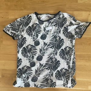 Vit t-shirt med svart palmtryck AOR - Vit t-shirt från AOR med svart tropiskt palm- och ananasmönster. Svarta kantband vid hals och ärmslut ger en snygg kontrast. Skön passform och perfekt för dig som gillar en chill och trendig stil.