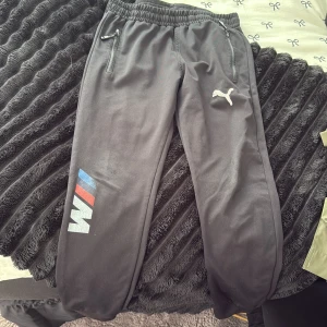 Svarta mjukisbyxor Puma x BMW M - Svarta mjukisbyxor från Puma med BMW M-logga i blått, rött och vitt på ena benet. Byxorna har resår i midjan, dragkedjefickor och Puma-logga broderad på låret. 