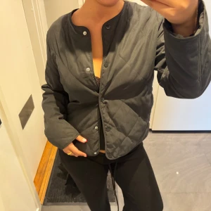 Svart quiltad jacka med tryckknappar - Säljer en svart quiltad jacka med rund halsringning och tryckknappar framtill. Jackan har en enkel, stilren design och är lätt vadderad. Perfekt för lager-på-lager och funkar till många olika outfits. Nyskick!