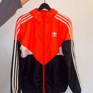 Adidas orange och mörkblå vindjacka S - Snygg vindjacka från Adidas i orange, mörkblå och vitt med klassiska tre ränder längs ärmarna. Jackan har huva med snörning, dragkedja framtill och meshfoder. Perfekt för dig som gillar sportig stil och streetwear.