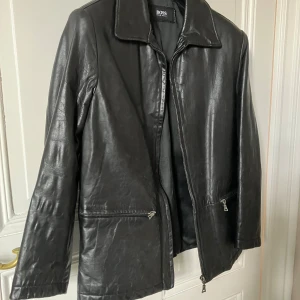 Svart skinnjacka från BOSS - Stilren svart skinnjacka från BOSS i 100% äkta läder. Jackan har klassisk krage, raka linjer och dragkedja framtill samt två snedställda fickor med dragkedja. Perfekt för dig som gillar en clean och tidlös look.