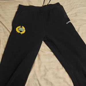 Svarta mjukisbyxor Craft Hammarby M - Svarta mjukisbyxor från Craft med Hammarby IF-logga på ena benet och broderad Craft-logga på andra. Byxorna har resår i midjan med snörning och är tillverkade i mjukt material, perfekta för chill eller träning.