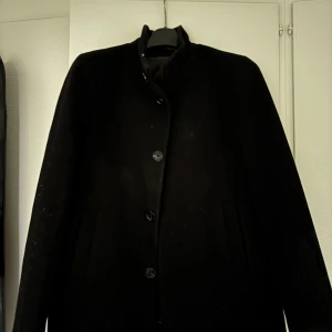 Svart rock från Matinique, stl S - Stilren svart kort rock från Matinique i klassisk modell med knäppning framtill och diskreta fickor. Jackan har en rak passform och hög krage, perfekt för dig som vill ha en clean och tidlös look.