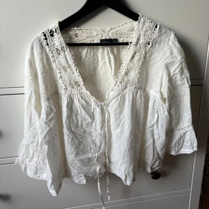 Vit spetsblus från Lindex XS/S - Romantisk vit blus från Lindex med v-ringning, virkade spetsdetaljer vid hals och axlar samt knyt framtill. Blusen har vida ärmar och är tillverkad i bomull med diskret prickstruktur. Perfekt för en bohemisk look.