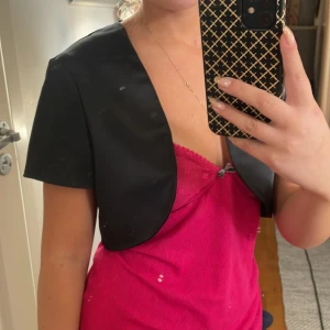 Svart kort väst i satin - Snygg svart kort väst i satin med enkel, stilren design och korta ärmar. Perfekt att slänga över en klänning eller topp för en extra touch. Västen har en öppen front och en elegant, minimalistisk look.