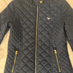 Quiltad svart jacka Hampton Republic - Snygg quiltad svart jacka från Hampton Republic med guldfärgade dragkedjor och ståkrage. Jackan har figurnära passform och två fickor med dragkedja. Perfekt för dig som gillar stilren och sportig look.