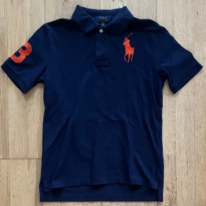 Marinblå piké från Polo Ralph Lauren - Marinblå pikétröja från Polo Ralph Lauren med orange broderad logga på bröstet. OBS! Storlek 10-12 men sitter som Xs. Tveka inte på att höra av dig om du har några frågor eller funderingar!