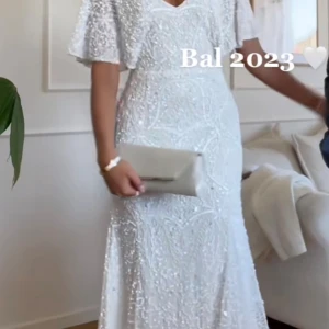 Vit glittrig långklänning med pärlor - Magisk vit långklänning med glittriga pärldetaljer och v-ringad hals. Klänningen har korta, vida ärmar och ett elegant fall. 