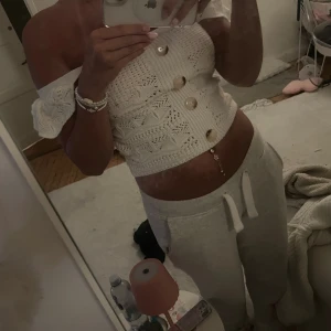 Vit stickad offshoulder topp med knappar - Supersöt vit stickad topp med offshoulder och stora dekorativa knappar framtill. Toppen har virkade detaljer och volang vid ärmarna, vilket ger en romantisk vibe. Perfekt för dig som gillar en feminin och trendig stil.