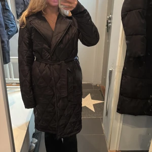 Svart quiltad kappa med bälte - Svart lång quiltad kappa från Zara med bälte i midjan och stora slag. Jackan har lång ärm och ett snyggt rutmönster. Perfekt för kyliga dagar och ger en clean och trendig look.