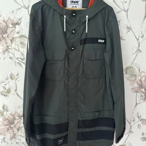 Skidjacka - Snygg grön vindjacka från Dope med svarta detaljer och vit dragkedja. Jackan har huva med snörning, stora fickor framtill och orange meshfoder på insidan. Perfekt för blåsiga dagar och streetwear-stil i skidbacken, Dope jacka 