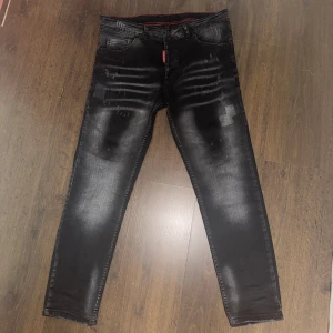 Svarta Dsquared2 jeans med slitningar - Snygga svarta jeans från Dsquared2 med slitna detaljer och tvättad look. Byxorna har smal passform, klassiska fem fickor och en röd broderad lönnlöv på bakfickan. Materialet är jeans och färgen är svart med grå tvättade partier.
