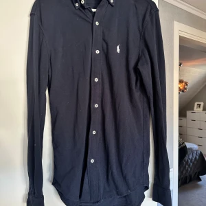 Mörkblå skjorta från Ralph Lauren XS - Klassisk mörkblå skjorta från Ralph Lauren i lätt mesh-bomull. Skjortan har button down-krage, vita knappar och den ikoniska broderade loggan på bröstet. Långärmad och perfekt för en clean och stilren look. Storleken är XS men sitter mer som S. Pris kan diskuteras vid snabb affär