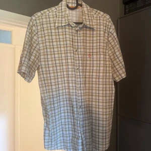 Rutig kortärmad skjorta från Fjällräven - Snygg rutig skjorta från Fjällräven i beige och grå toner. Skjortan har korta ärmar, klassisk krage och knappar hela vägen. På bröstet finns en diskret ficka med Fjällrävens röda logotyp. Perfekt för dig som gillar friluftsstil och chill vibe.