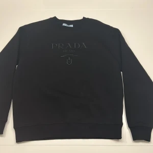 Svart sweatshirt från Prada Milano - Svart sweatshirt från Prada Milano med broderad logga framtill. Klassisk rund halsringning, ribbade muddar och mjukt material i bomullsblandning. Perfekt för dig som gillar stilrena designer och exklusiva märken. Använd kanske 1-3 gånger 