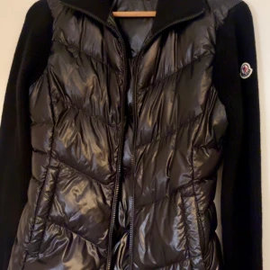 Svart pufferjacka från Moncler - Snygg svart pufferjacka från Moncler med glansig, quiltad framsida och stickade ärmar. Jackan har dragkedja framtill och Moncler-logga på ärmen. Perfekt för dig som vill ha en stilren och sportig look.