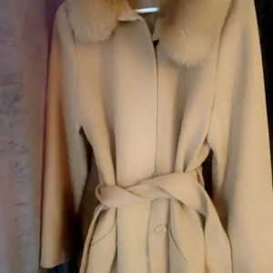 Beige kappa med fuskpäls från JACKIE - Stilren beige kappa med bred krage i fuskpäls och knytskärp i midjan. Ny pris 6000. Klassisk rak modell med fickor framtill och dold knäppning. Perfekt för dig som vill ha en elegant och tidlös look.