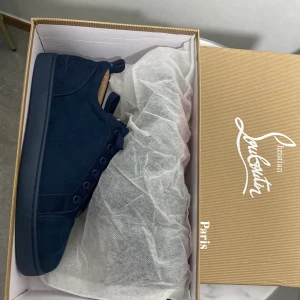 Mörkblå sneakers från Christian Louboutin - Snygga mörkblå sneakers från Christian Louboutin i mocka med klassisk röd sula. Skorna har rund tå, snörning och diskret broderad logga på sidan. Insidan är ljusbeige och de levereras med originalkartong och dustbag. Perfekta för dig som vill sticka ut med stil.