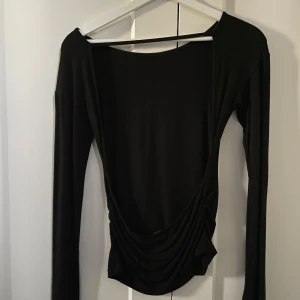 Svart långärmad topp med öppen rygg - Säljer en svart långärmad topp med en djup öppen rygg och snygg drapering nedtill. Toppen har en tight passform och är tillverkad i ett mjukt, stretchigt material som sitter skönt på kroppen. 