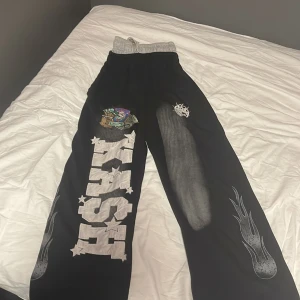 Svarta cargopants med tryck och snörning - Unika svarta cargopants med bred passform, elastisk midja med grå snörning och stora fickor bak. Byxorna har coola tryck med texten 'KASH', stjärnor, eldflamma och färgglada detaljer på benen. Perfekta för dig som vill sticka ut med streetstyle. Double waisted