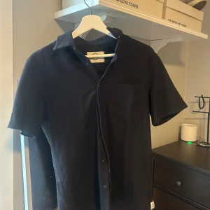 Svart kortärmad skjorta GOODS S - Svart kortärmad skjorta från GOODS Surfside Supply. Skjortan har klassisk krage, knäppning framtill och en bröstficka. Perfekt för en clean och avslappnad stil. Tillverkad i mjuk bomull för skön känsla hela dagen. Priset går alltid att diskutera!