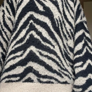 Zebramönstrad stickad tröja från Ellos - Stickad tröja från Ellos med svart och beige zebramönster. Tröjan har rund halsringning, ribbade muddar och är långärmad. Perfekt för dig som vill sticka ut med ett djurmönster och gillar en mysig, lite oversized känsla.
