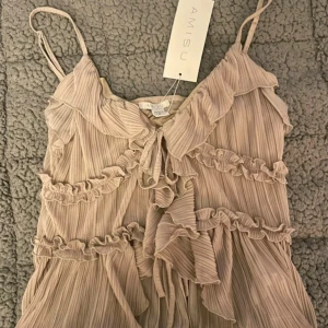 Beige volangtop från Amisu - Supersöt beige linnetopp från Amisu med tunna axelband, volangdetaljer och plisserat tyg. Toppen har knyt framtill och en luftig, flowy känsla som passar perfekt till sommaren. Perfekt för dig som gillar romantisk och feminin stil.