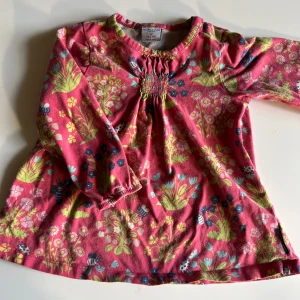 Blommig långärmad tröja Polarn O. Pyret - Supersöt långärmad tröja från Polarn O. Pyret i rosa med färgglatt blommönster och små bin. Tröjan har rynkdetalj vid halsen och är tillverkad i mjuk bomull. Perfekt för vår och sommar när man vill ha något lekfullt och bekvämt.