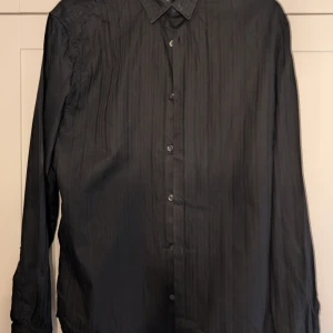 Svart randig skjorta från H&M L - Svart skjorta från H&M i storlek L med diskreta vertikala ränder. Klassisk krage, knäppning framtill och långa ärmar.